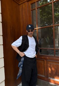 Veste noire sans manches sur une chemise blanche, pantalon noir et bandana bleu. Accessoires : lunettes de soleil et casquette noire avec un logo. Fond en bois.