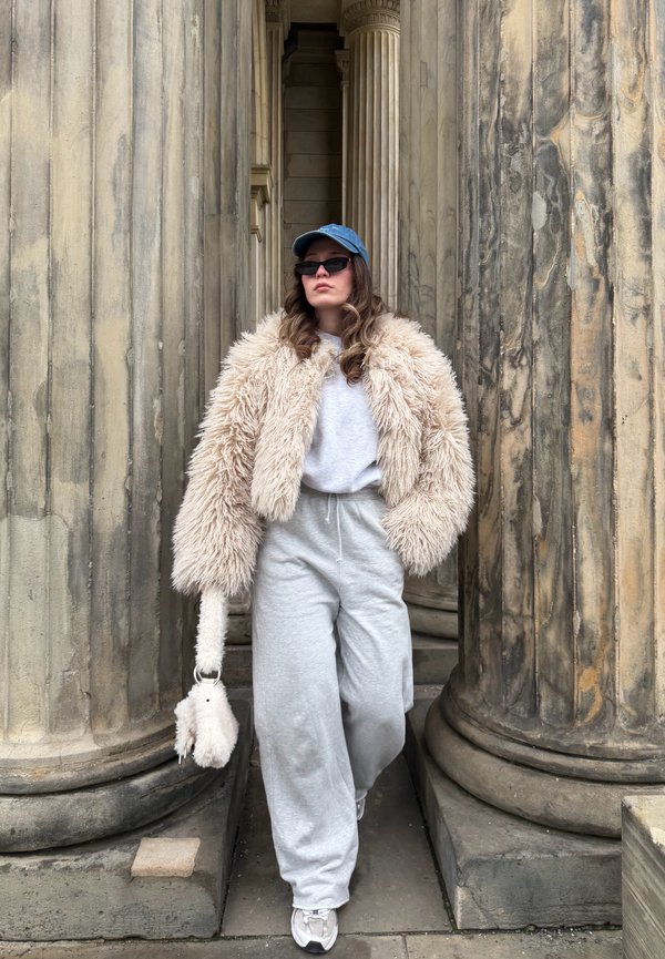 Woman wearing fluffy beige jacket, gray sweatpants, white sneakers, blue cap, and black sunglasses walking between large stone columns.