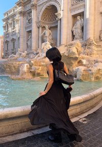 Γυναίκα με μαύρο φόρεμα και μπότες που κρατά τσάντα δίπλα σε ιστορικό περίτεχνο σιντριβάνι με αγάλματα και τιρκουάζ νερό στο φως της ημέρας.