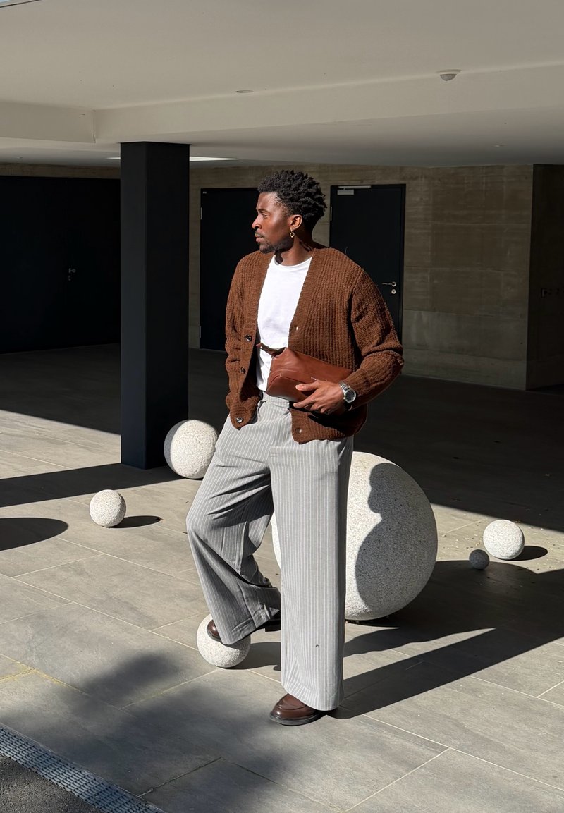 Homme portant un cardigan marron, une chemise blanche et un pantalon gris à fines rayures, debout à l'intérieur près de grandes sphères texturées à la lumière du soleil et à l'ombre.