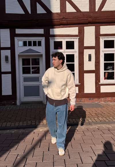 Joven con sudadera beige y vaqueros azules caminando por una calle empedrada iluminada por el sol, frente a un edificio entramado con ventanas blancas.