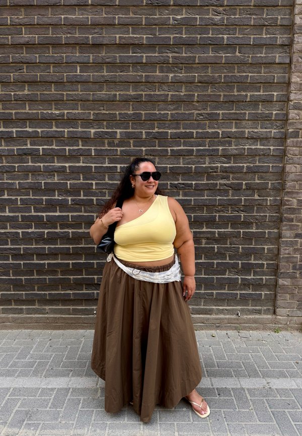 Femme debout près d'un mur en briques, portant des lunettes de soleil, un haut jaune à une épaule, une jupe longue marron, des sandales, et tenant un sac noir sur son épaule.