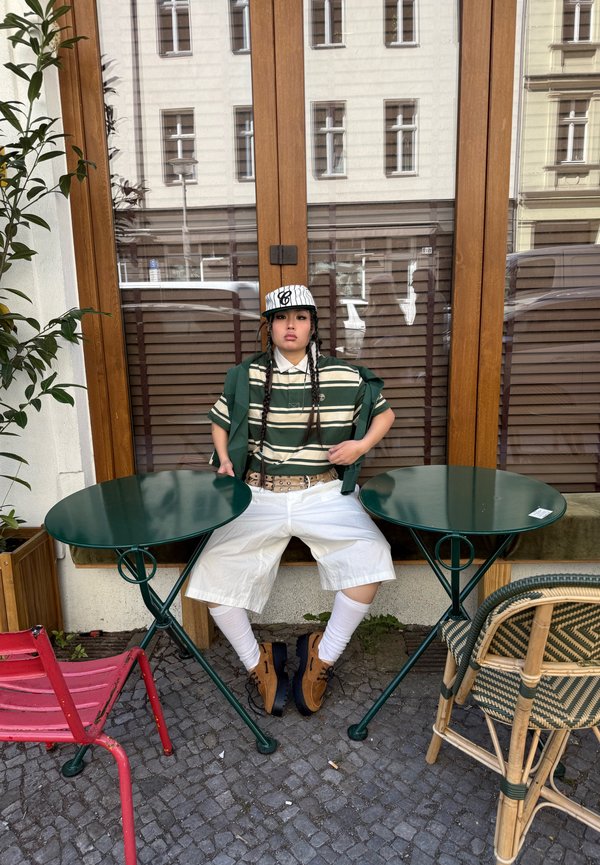 Giovane che indossa una maglietta a righe, pantaloncini bianchi, cappellino e scarpe marroni, seduto tra due tavoli rotondi verdi fuori da un bar.