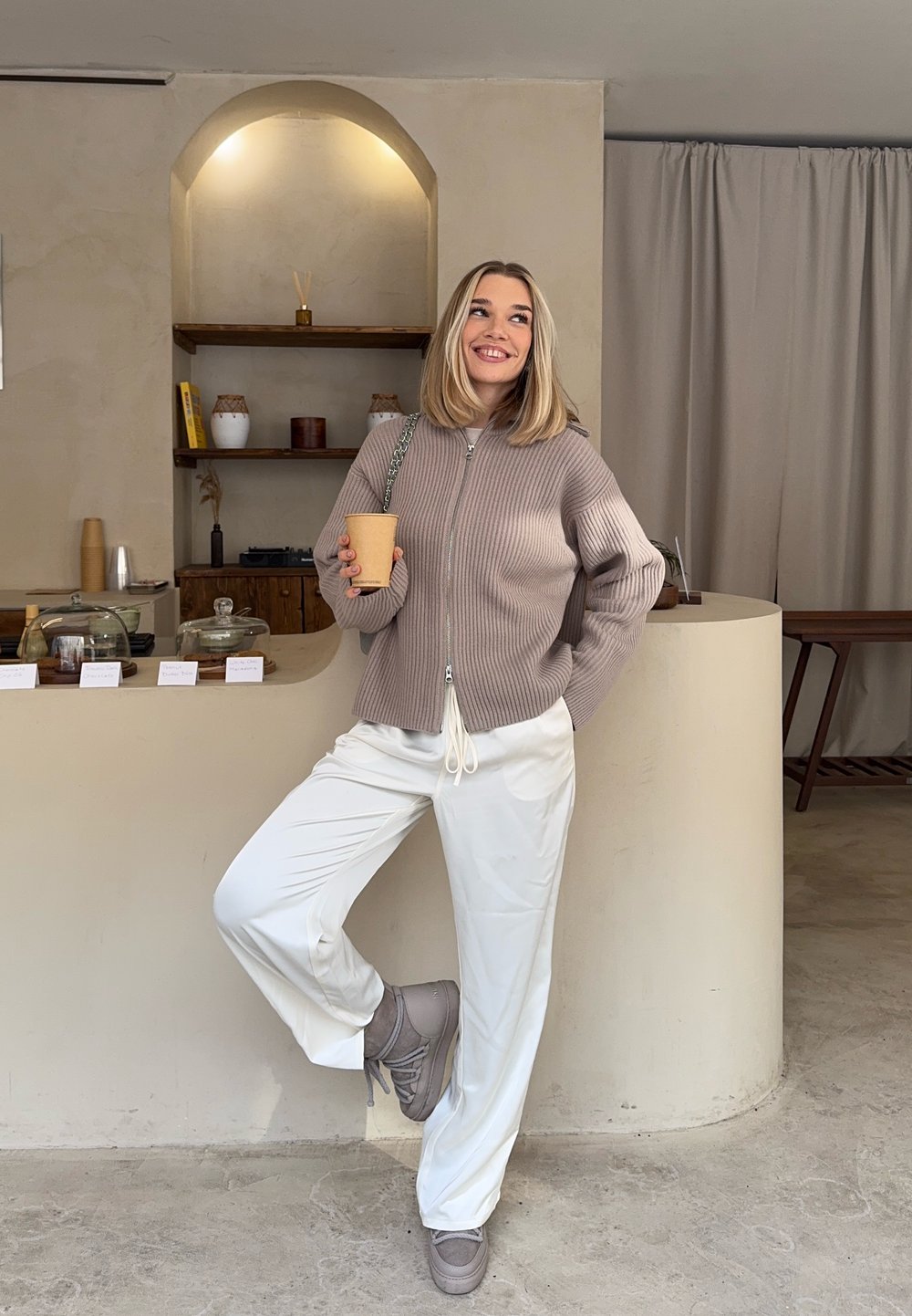 Jeune femme en pull à fermeture éclair beige et pantalon blanc tenant un gobelet en papier, appuyée contre un comptoir incurvé dans un intérieur de café minimaliste.