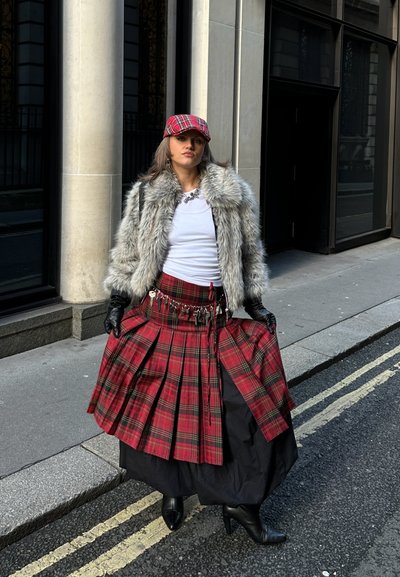 Kilt de tartán rojo con pliegues, enagua negra, abrigo de piel sintética, guantes negros y gorra a juego de tartán rojo. Fondo de calle urbana.