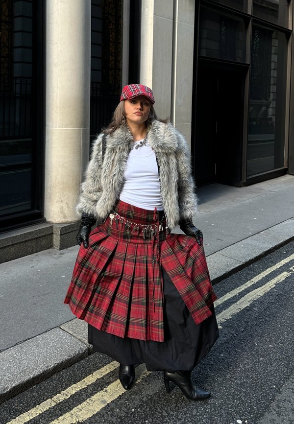 Punaruudullinen pliseerattu kilttohousu, musta alushame, tekoturkistakki, mustat käsineet ja yhteensopiva punaruudullinen lakki. Kaupungin katuympäristö taustalla.