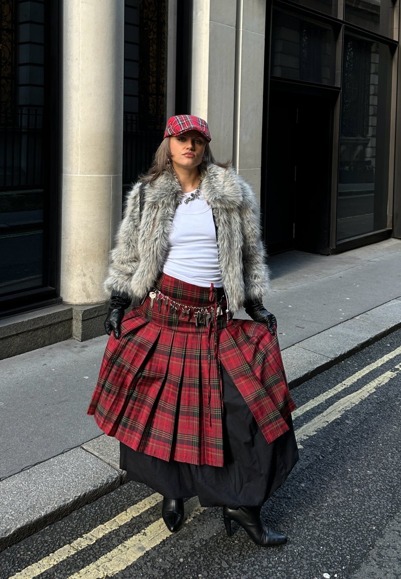 Kilt écossais à carreaux rouges avec plis, jupon noir, manteau en fausse fourrure, gants noirs et casquette assortie à carreaux rouges. Décor urbain en arrière-plan.