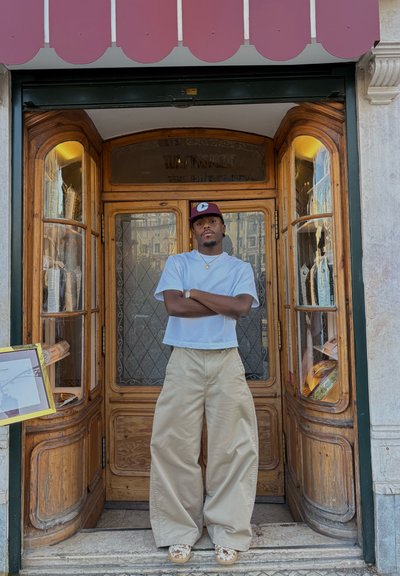 Hombre con gorra burdeos, camiseta blanca, pantalones beige de pierna ancha y zapatos estampados, de pie con los brazos cruzados en la puerta de entrada de una tienda de madera.