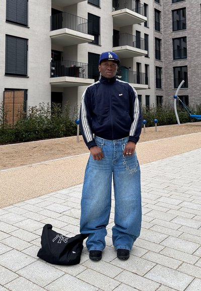 Chaqueta navy de cremallera con rayas blancas, jeans azules de corte amplio, zapatos negros y una gorra azul. Una bolsa de mano negra descansa en el suelo.
