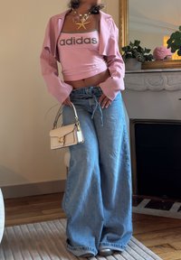 Persona con top corto rosa de Adidas, chaqueta rosa, jeans anchos azules, sosteniendo un pequeño bolso blanco, de pie en interiores cerca de una chimenea y un espejo.