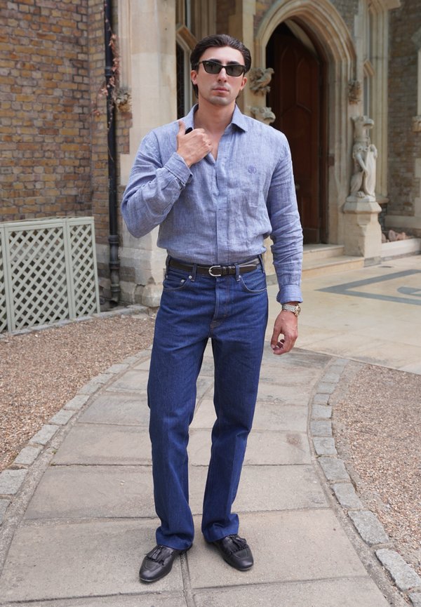 Homme portant des lunettes de soleil, une chemise bleu clair, un jean bleu foncé et des chaussures noires, debout sur un chemin en pierre devant un bâtiment historique.
