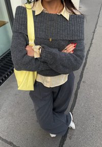 Grauer gestrickter Pullover mit Rippdesign, über einem gelb gestreiften Hemd getragen. Begleitet von einer gelben Tasche und weiten grauen Hosen. Rote Nägel sichtbar.