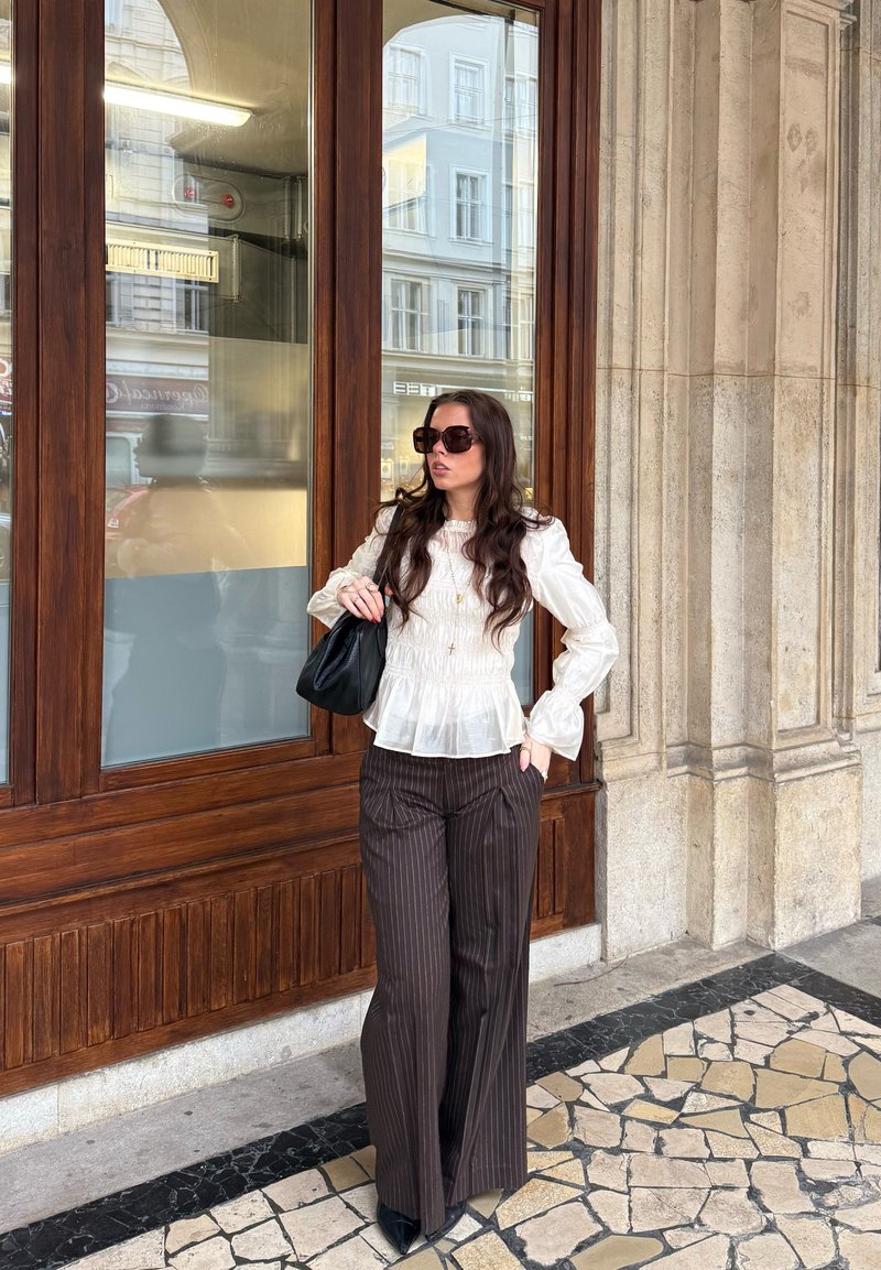 Γυναίκα με μακριά σκούρα μαλλιά και γυαλιά ηλίου, που φοράει άσπρο πουκάμισο και παντελόνι με ρίγες, κρατώντας μαύρη τσάντα, στέκεται δίπλα σε ένα ξύλινο παράθυρο.