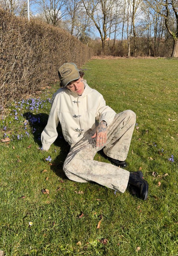 Person trägt eine beige Jacke, gemusterte Hosen, schwarze Stiefel und eine Kappe sitzt auf dem Gras neben einer Hecke mit violetten Blumen unter kahlen Bäumen.