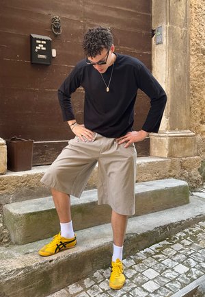 Camisa negra de manga larga, pantalones cortos beige con pliegues, calcetines blancos y zapatillas amarillas con acentos negros. De pie en los escalones de piedra contra un fondo rústico.