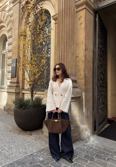 Abrigo de color crema de doble botonadura con mangas anchas, vaqueros acampanados de color azul oscuro, sosteniendo un bolso de ante marrón. Pared de piedra y planta de fondo.