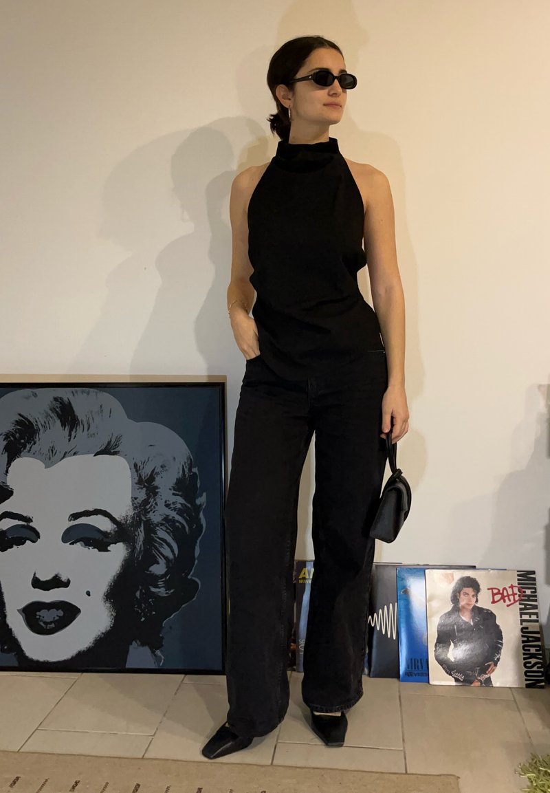 Femme portant un haut noir sans manches, un pantalon, des lunettes de soleil, tenant un petit sac ; une affiche encadrée de Marilyn Monroe et des disques vinyles posés sur le sol à proximité.
