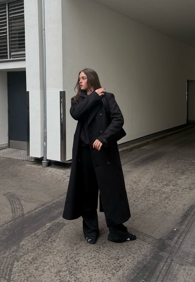 Abrigo largo negro con diseño de doble botonadura, combinado con pantalones y zapatos negros a juego. Tejido texturizado, estilo minimalista.