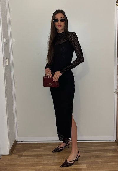 Mujer con vestido de encaje negro, gafas de sol oscuras, sosteniendo un bolso burdeos, de pie sobre un suelo de madera frente a una puerta blanca.