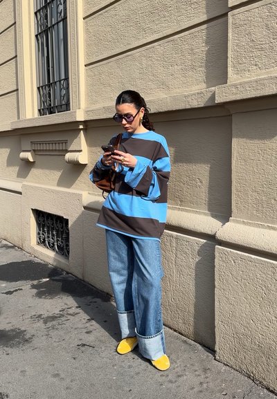 Top de rayas de gran tamaño en azul y marrón, jeans de pierna ancha de mezclilla con puños en azul claro y zapatos amarillos de deslizar, de pie frente a una pared texturizada.