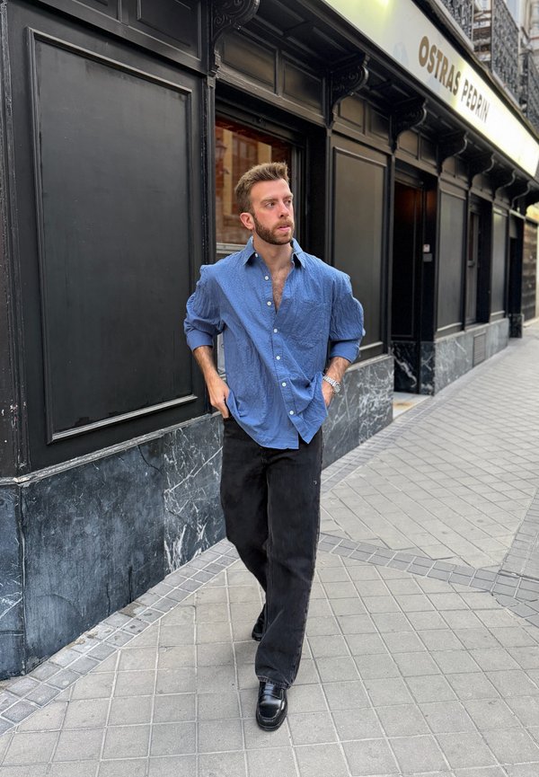 Homme en chemise bleue et pantalon noir marchant sur un trottoir carrelé près d'un bâtiment noir avec un panneau indiquant Ostras Pedrin.