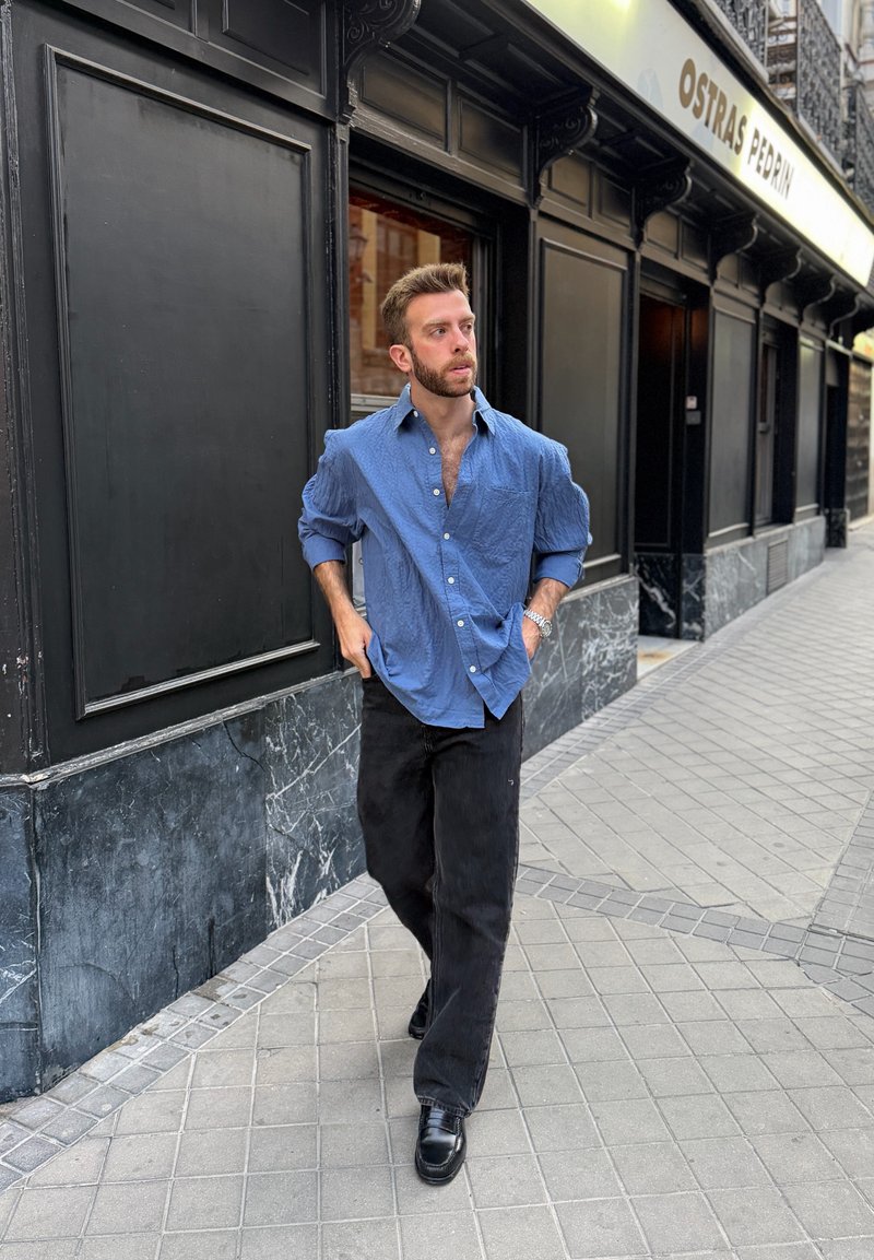 Homme en chemise bleue et pantalon noir marchant sur un trottoir carrelé près d'un bâtiment noir avec un panneau indiquant Ostras Pedrin.