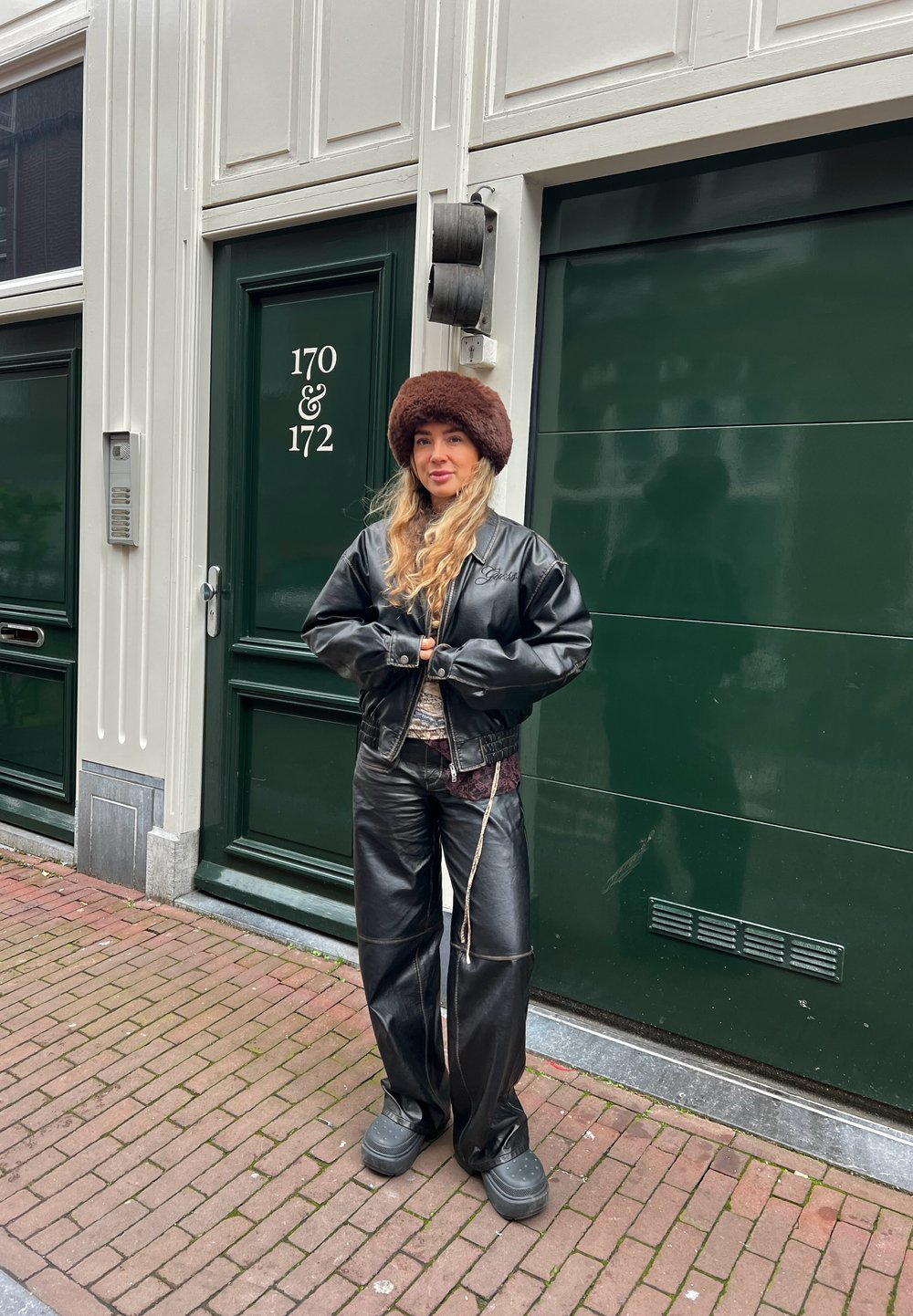 Femme portant un chapeau marron en fourrure, une veste en cuir noire et un pantalon, debout sur un trottoir en briques devant des portes vertes numérotées 170 et 172.