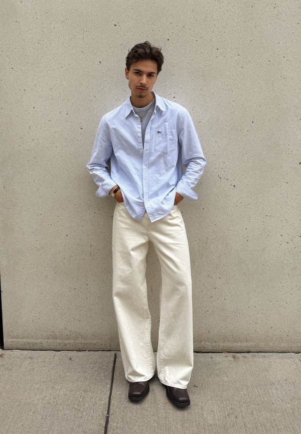 Jeune homme debout contre un mur uni portant une chemise bleu clair, un sous-vêtement gris, un pantalon large blanc et des chaussures marron foncé avec les mains dans les poches.