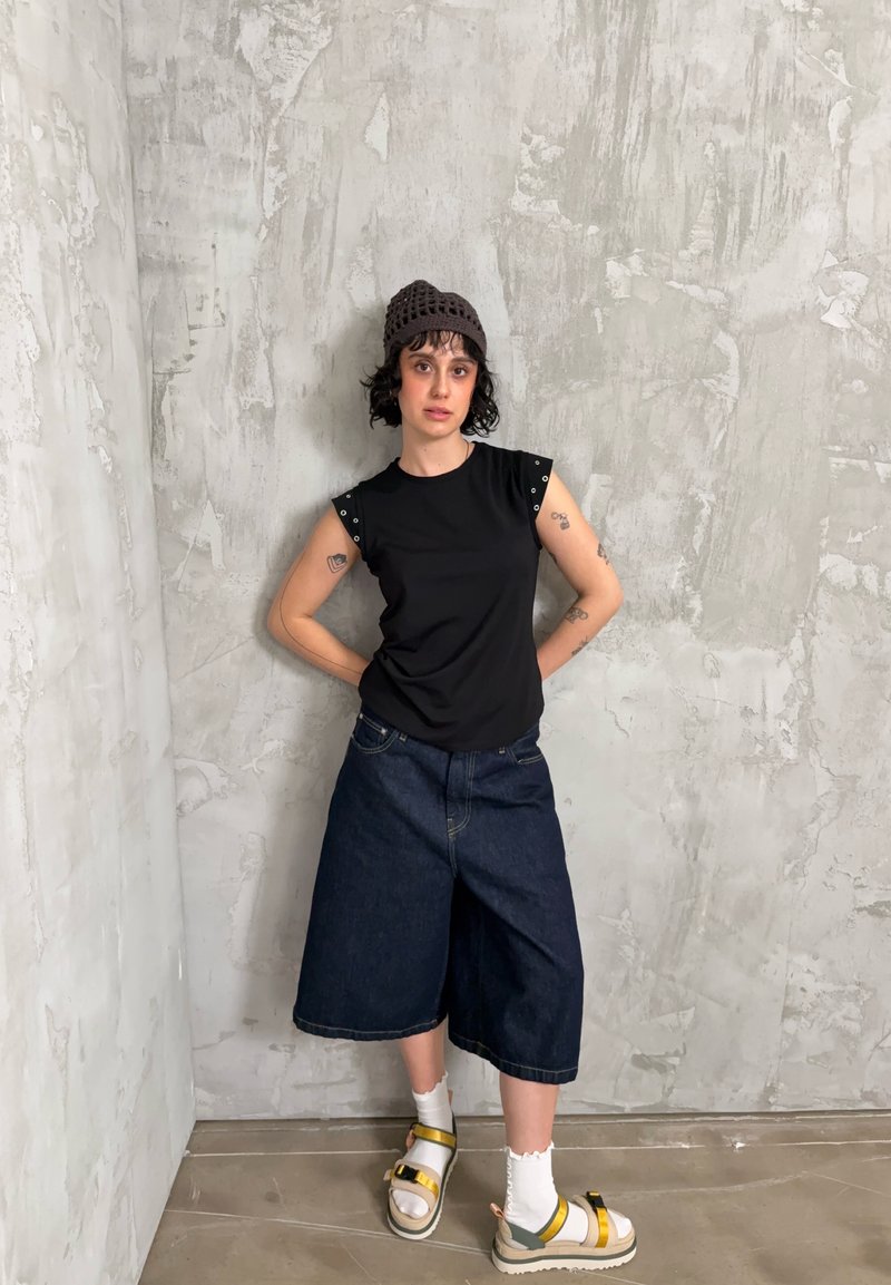 Personne aux cheveux bouclés portant un haut noir sans manches, un short en jean large et foncé, des chaussettes blanches et des sandales jaunes à brides, debout devant un mur gris texturé.