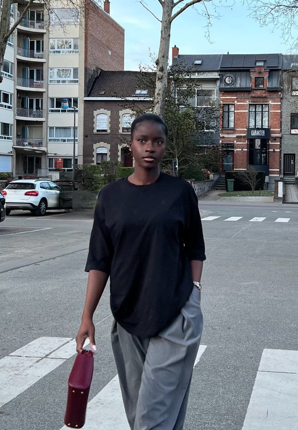 Joven mujer con camisa negra y pantalones grises camina por una calle urbana sosteniendo un bolso de mano color granate, con edificios y coches de fondo.