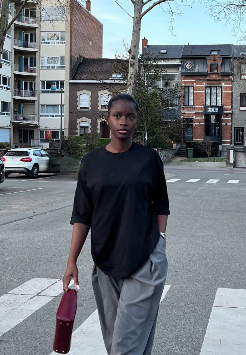 Giovane donna con maglietta nera e pantaloni grigi attraversa una strada urbana tenendo una pochette color bordeaux, con edifici e auto sullo sfondo.