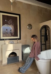 Hvid fritstående badekar ved siden af en stenpejs, indrammet portræt af en ældre mand, blåstribet lænestol, teksturerede beige vægge.