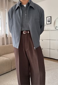 Pilkų sagų švarkas su apykakle, derinamas su tamsiai pilku polo marškinėliu ir rudos spalvos raukšlėtomis kelnėmis. Rudas diržas ir neutralios spalvos fonas.