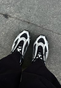 Tênis atléticos pretos e prateados com parte superior em malha, design ondulado único e detalhes estampados. Exibidos em pavimento cinza.