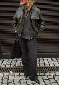 Veste en cuir vert olive avec des patchs aux épaules, pull gris, sac fourre-tout tissé noir, jean foncé et talons pointus marron. Fond en pavé de pierre plate.