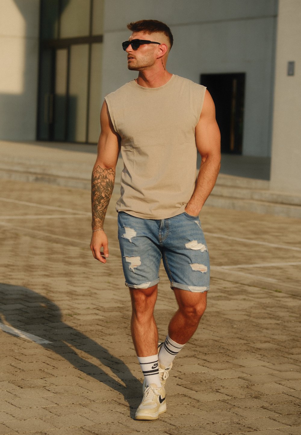 Beige ermeløs topp, slitte denimshorts, hvite Nike-sneakers, svart-hvite stripete sokker og sorte solbriller. Brun hud og tatovering på armen.