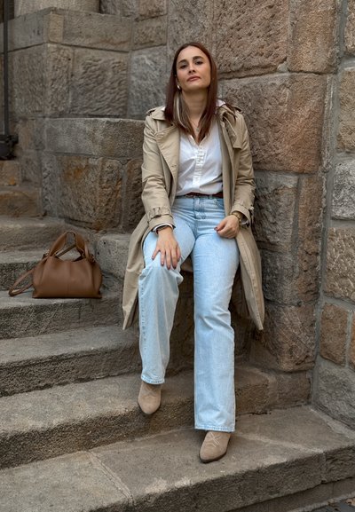 Gabardina beige con un diseño estructurado, blusa blanca, jeans acampanados de color azul claro y botines beige. Cartera marrón colocada al lado.