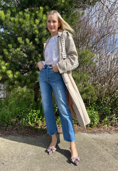 Trench beige con acentos de tachuelas, blusa blanca, jeans azules de cintura alta y tacones con estampado de leopardo. Follaje verde en el fondo.