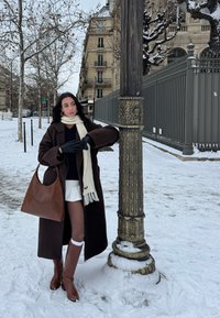 Femme en manteau marron, écharpe blanche et bottes, debout près d'un réverbère orné dans une rue urbaine enneigée avec des arbres dénudés et des bâtiments historiques.