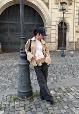 Joven con chaqueta beige, camisa blanca y gorra negra apoyándose en un farol en una calle empedrada con un edificio histórico de fondo.