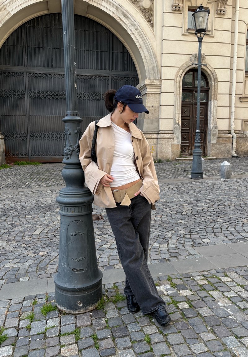 Jeune personne en veste beige, chemise blanche et casquette noire appuyée contre un lampadaire dans une rue pavée avec un bâtiment historique en arrière-plan.