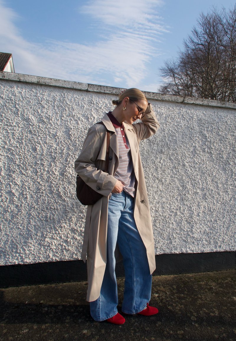 Mulher vestindo casaco trench bege, camisa polo às riscas, jeans de pernas largas, sapatos vermelhos e óculos de sol, em pé junto a uma parede branca texturizada ao ar livre.