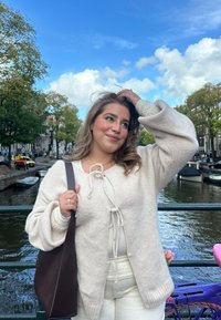 Ljust beige stickad cardigan med knytning, kombinerad med en ljus topp och vita byxor. Mörkbrun axelväska. Utomhus vid en kanal.