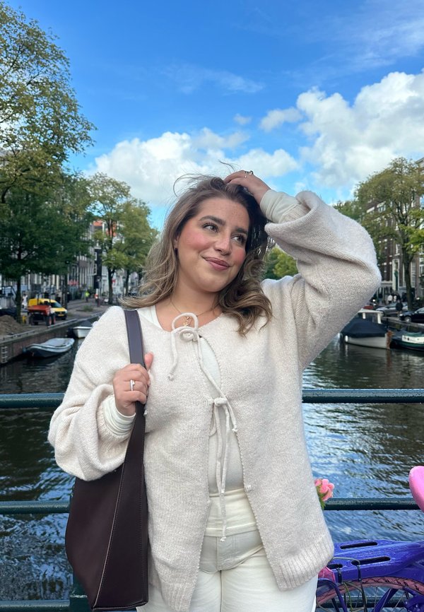 Cardigan lavorato a maglia beige chiaro con chiusura a fiocco, abbinato a una maglietta leggera e pantaloni bianchi. Borsa a spalla marrone scuro. Ambientazione all'aperto lungo un canale.