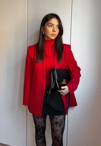Czerwony żakiet na czerwonym golfie, czarna spódnica i wzorzyste czarne rajstopy; trzymając małą czarną torebkę z złotym zapięciem.