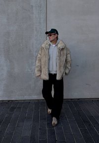 Crèmekleurige jas van nepbont met een grote kraag, gedragen over een grijze hoodie en zwarte broek. Gecombineerd met beige sneakers en een groene pet.