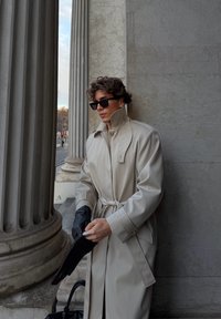 Trench-coat beige à col montant, avec un lien sur le côté et des coutures détaillées. Des gants en cuir noir sont enfilés. Mur en pierre en arrière-plan.