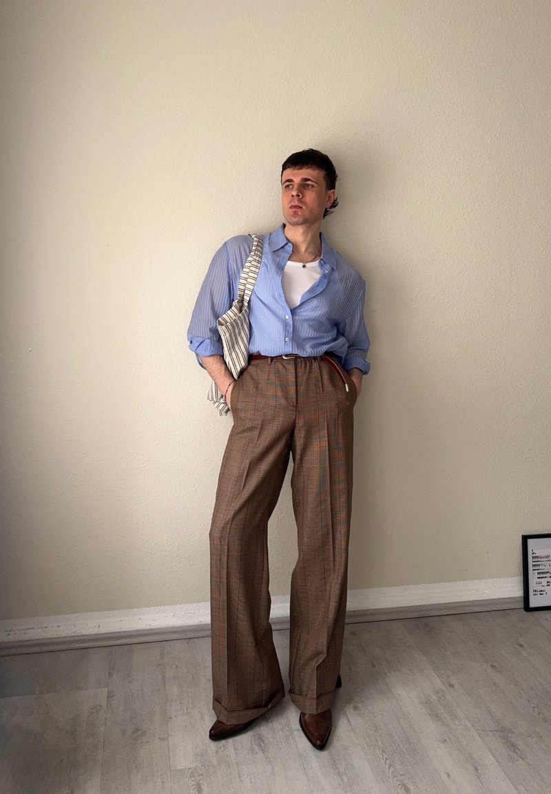 Person trägt ein blau gestreiftes Hemd über einem weißen Top, braune karierte weite Hosen und braune spitze Schuhe, steht vor einer beigen Wand mit Tragetasche.
