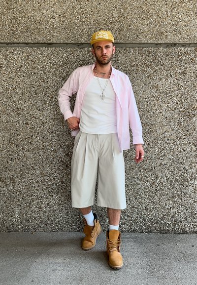 Persona que lleva una camisa a rayas rosas, una camiseta blanca sin mangas, pantalones cortos plegados beige, una gorra amarilla y botas de trabajo marrones con calcetines blancos. Fondo texturizado.