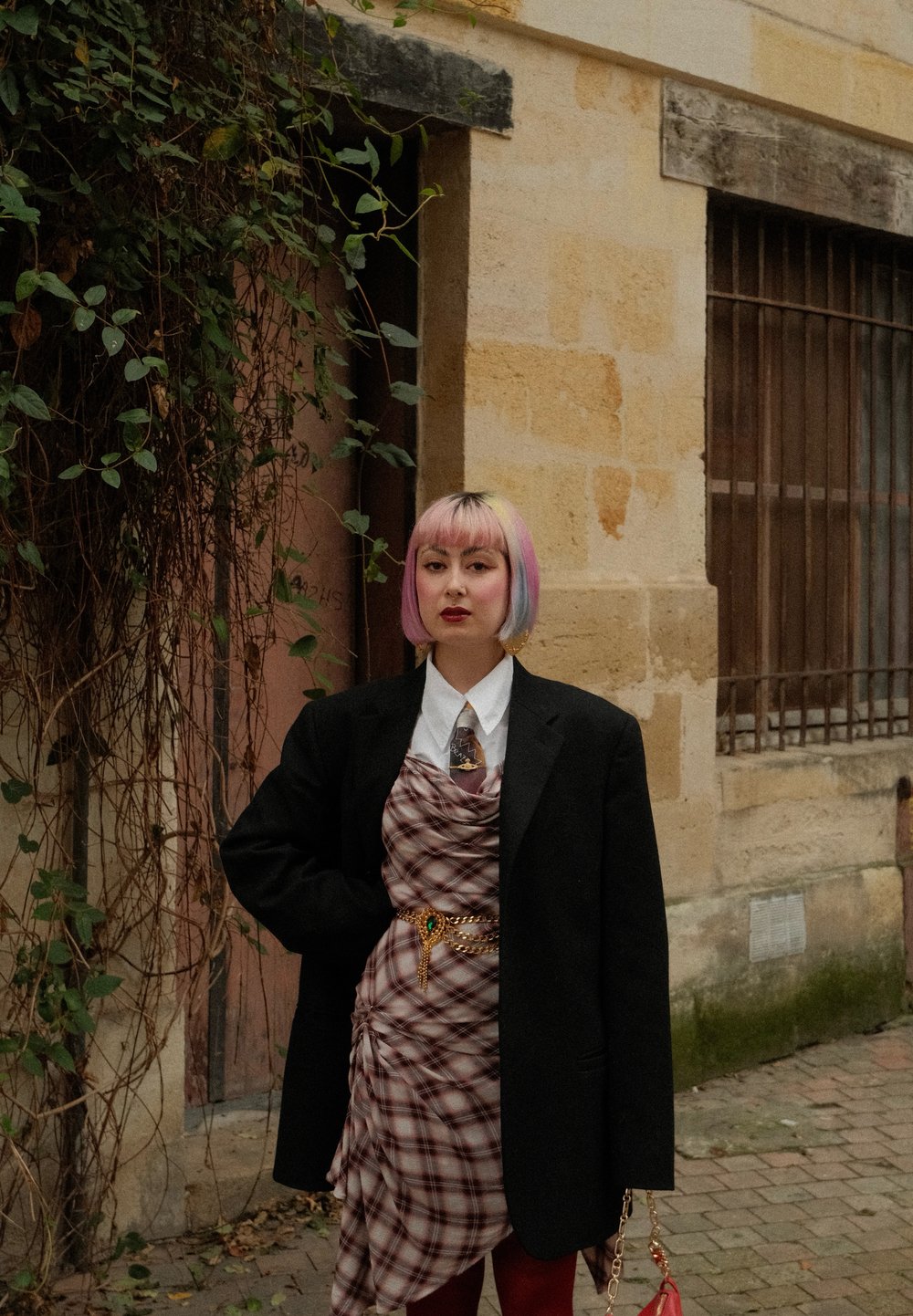Rutet kjole draperet over en hvid skjorte, lagdelt med en sort oversized blazer. Har et guld kædebelte og levende pink hår.